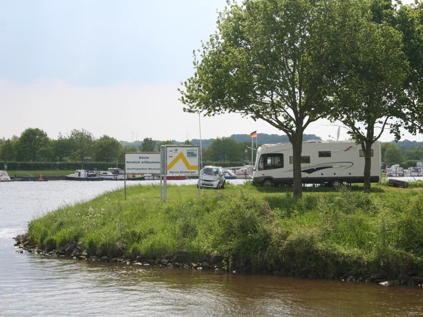 Wohnmobilstellplatz Marina Mehlbergen Wohnmobil auf begrüntem Platz an einer Marina, mit Bäumen und ruhigem Wasser im Hintergrund.