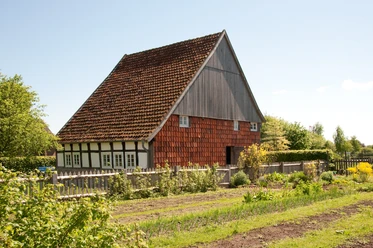 Fachwerkhaus mit roten Holzschindeln, umgeben von einem Garten und grüner Landschaft unter klarem Himmel.