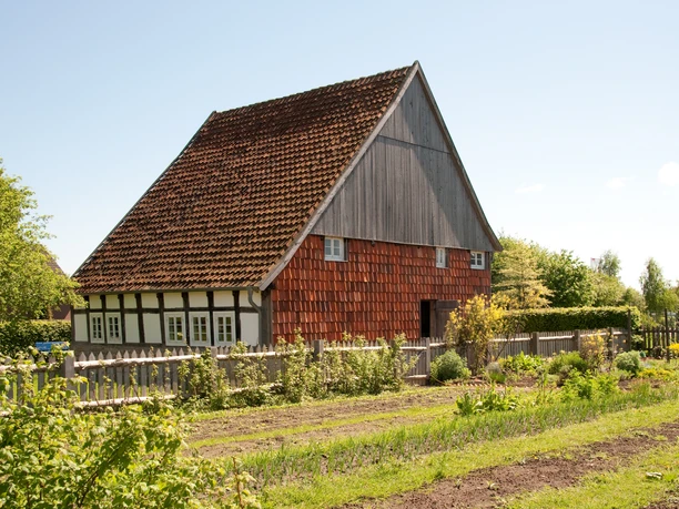 Fachwerkhaus mit roten Holzschindeln, umgeben von einem Garten und grüner Landschaft unter klarem Himmel.