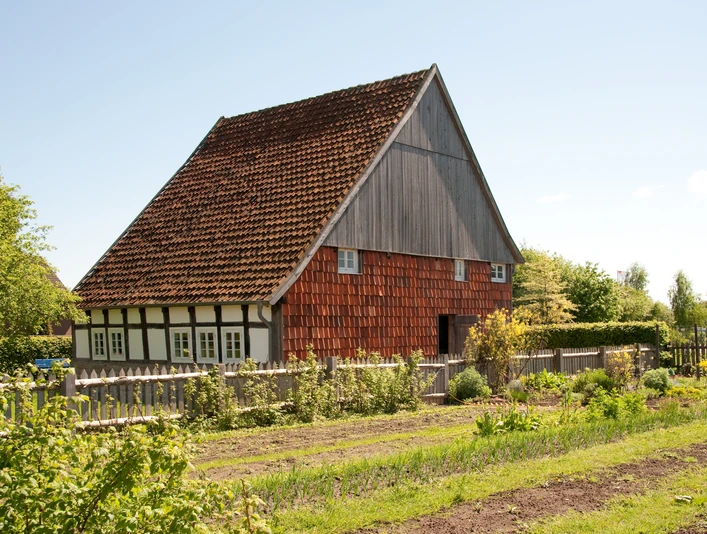LWL-Industriemuseum Lage: Kotten Fachwerkhaus mit roten Holzschindeln, umgeben von einem Garten und grüner Landschaft unter klarem Himmel.