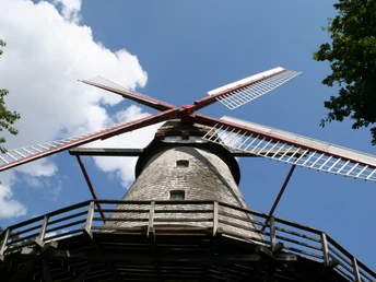 Historische Windmühle mit rot-weißen Flügeln und Holzverkleidung vor blauem Himmel mit Wolken.