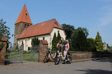 Two cyclists in conversation in front of the historic Eystrup church with its striking tower.