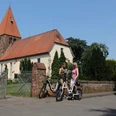 Zwei Fahrradfahrerinnen im Gespräch vor der historischen Kirche Eystrup mit markantem Turm.