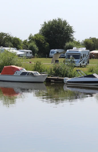 Wohnmobile und Boote am idyllischen Flussufer, umgeben von Bäumen und grünen Wiesen.