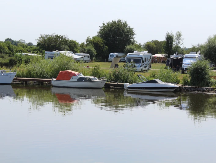 Wohnmobile und Boote am idyllischen Flussufer, umgeben von Bäumen und grünen Wiesen.