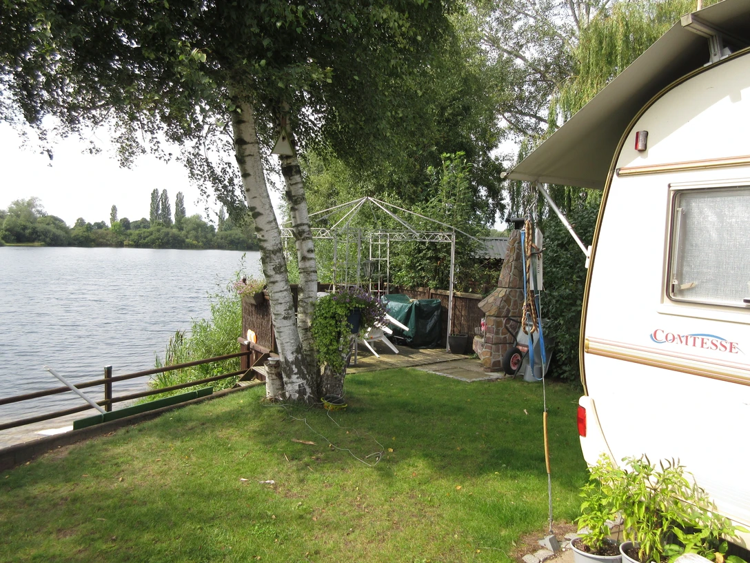 Grüne Wiese mit Wohnwagen am Seeufer, umgeben von Bäumen und einem Pavillon im Hintergrund.
