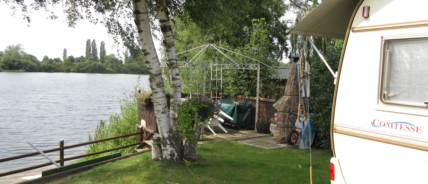 Grüne Wiese mit Wohnwagen am Seeufer, umgeben von Bäumen und einem Pavillon im Hintergrund.