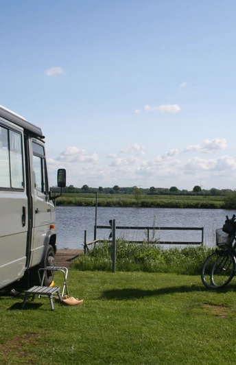 Wohnmobil steht auf einer Wiese am See, neben zwei Fahrrädern und einem Stuhl mit Blick aufs Wasser.