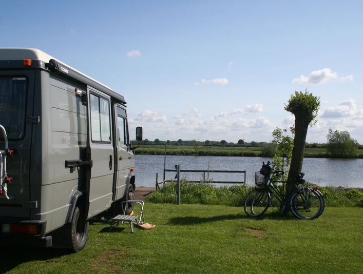 Campingplatz am See Wohnmobil steht auf einer Wiese am See, neben zwei Fahrrädern und einem Stuhl mit Blick aufs Wasser.Motorhome parked on a meadow by the lake, next to two bicycles and a chair with a view of the water.Autocamper parkeret på en eng ved søen, ved siden af to cykler og en stol med udsigt over vandet.Camper geparkeerd op een weiland bij het meer, naast twee fietsen en een stoel met uitzicht op het water.