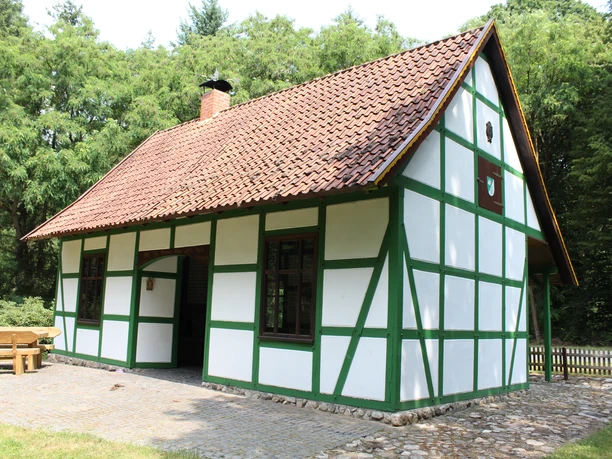 White half-timbered house with green beams, red tiled roof, surrounded by trees and a gravel path.