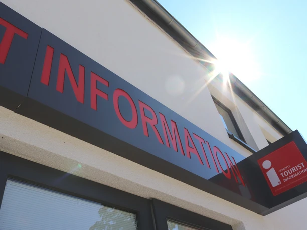 Eingang einer Tourist-Information mit rotem Schriftzug, Sonnenschein reflektiert am Gebäude.