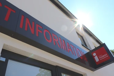 Tourist-Information Bad Lippspringe Eingang einer Tourist-Information mit rotem Schriftzug, Sonnenschein reflektiert am Gebäude.