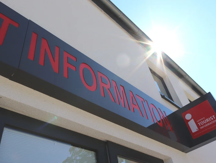 Tourist-Information Bad Lippspringe Eingang einer Tourist-Information mit rotem Schriftzug, Sonnenschein reflektiert am Gebäude.