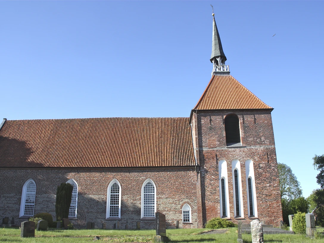 Evangelisch-reformierte Kirche in Rysum Backsteinkirche mit hohem Turm und spitzem Helm, umgeben von alten Grabsteinen unter blauem Himmel.
