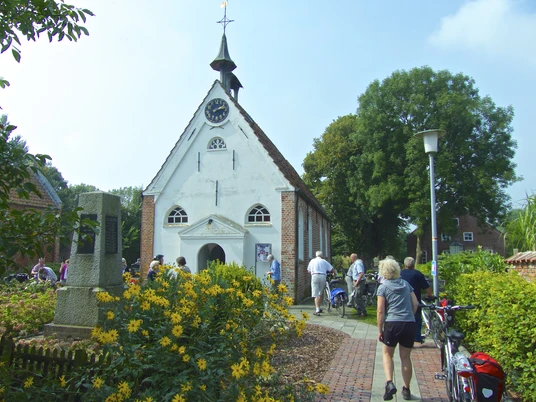 Kirche Woltzeten Weiße Backsteinkirche mit Uhrturm und Garten, Menschen mit Fahrrädern gehen zum Eingang.