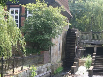 Wassermühle Sudweyhe Alte Backstein-Wassermühle mit hölzernem Rad, flankiert von Bäumen und ruhigem Bachlauf.