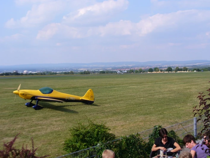 Gelbes Sportflugzeug auf einer grünen Landebahn des Flugplatzes Paderborn-Haxterberg. Im Hintergrund erstreckt sich eine weite Landschaft unter wolkigem Himmel.
