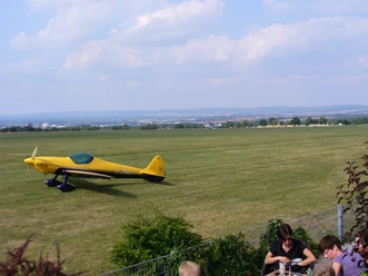 Flugplatz Paderborn-Haxterberg (Landebahn) Gelbes Sportflugzeug auf einer grünen Landebahn des Flugplatzes Paderborn-Haxterberg. Im Hintergrund erstreckt sich eine weite Landschaft unter wolkigem Himmel.