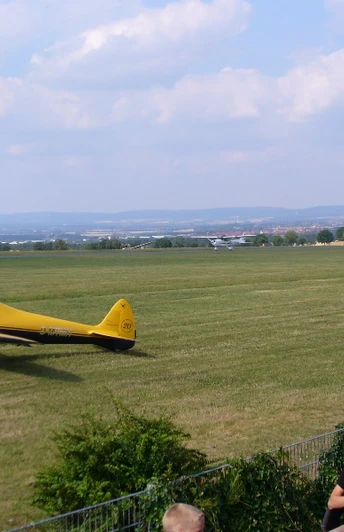 Flugplatz Paderborn-Haxterberg (Landebahn) Gelbes Sportflugzeug auf einer grünen Landebahn des Flugplatzes Paderborn-Haxterberg. Im Hintergrund erstreckt sich eine weite Landschaft unter wolkigem Himmel.