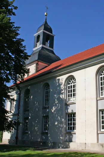 Eine Kirche mit rotem Ziegeldach und weißer Fassade, umgeben von Bäumen und blauem Himmel.