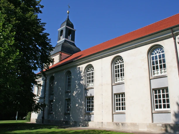 A church with a red tiled roof and white façade, surrounded by trees and a blue sky.