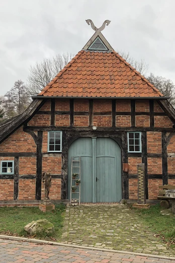 Altes Fachwerkhaus mit rotem Ziegeldach und blauer Holztür, umgeben von Bäumen und Infotafeln.