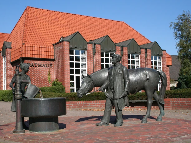 Een bronzen beeld van een vrouw met een paard voor een bakstenen stadhuis met een rood pannendak.