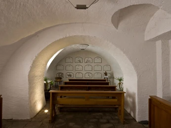 Barocke Gruft mit schlichten weiß-grauen Wänden, Holzbänken und historischen Grabplatten.