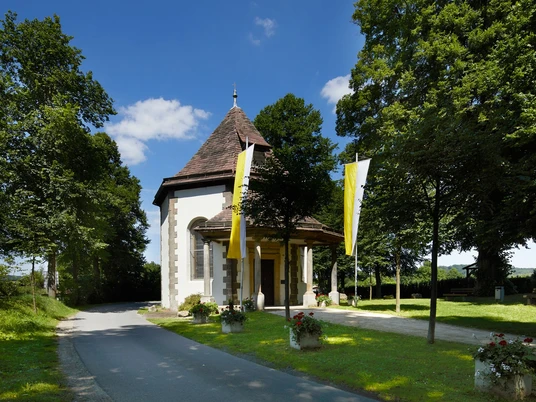 Annenkapelle mit spitzem Holzdach und gelben Fahnen, umgeben von Bäumen, neben einer Straße.