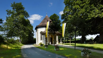 Annenkapelle Annenkapelle mit spitzem Holzdach und gelben Fahnen, umgeben von Bäumen, neben einer Straße.