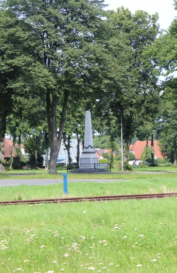 Eine grüne Wiese mit Eisenbahnschienen und einem markanten Denkmal, umgeben von hohen Bäumen.