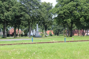 Denkmal auf dem Marktplatz Eine grüne Wiese mit Eisenbahnschienen und einem markanten Denkmal, umgeben von hohen Bäumen.