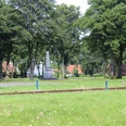 Denkmal auf dem Marktplatz Eine grüne Wiese mit Eisenbahnschienen und einem markanten Denkmal, umgeben von hohen Bäumen.
