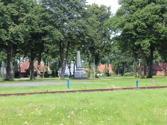 Denkmal auf dem Marktplatz Eine grüne Wiese mit Eisenbahnschienen und einem markanten Denkmal, umgeben von hohen Bäumen.