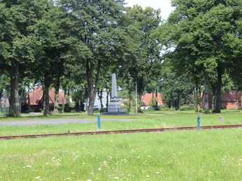 Denkmal auf dem Marktplatz Eine grüne Wiese mit Eisenbahnschienen und einem markanten Denkmal, umgeben von hohen Bäumen.