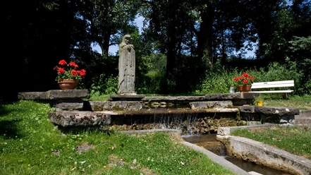 Die Statue der Mutter-Gottes steht umgeben von leuchtenden Geranien an einem kleinen Wasserlauf.