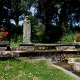 Die Statue der Mutter-Gottes steht umgeben von leuchtenden Geranien an einem kleinen Wasserlauf.
