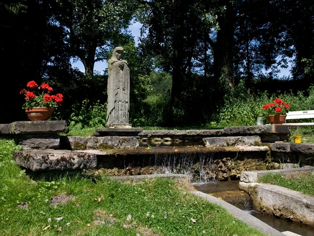 Die Statue der Mutter-Gottes steht umgeben von leuchtenden Geranien an einem kleinen Wasserlauf.