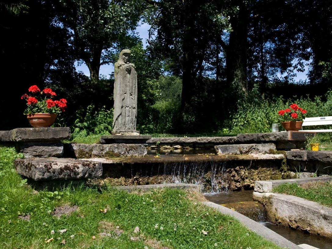 Mutter-Gottes-im-Wasser Die Statue der Mutter-Gottes steht umgeben von leuchtenden Geranien an einem kleinen Wasserlauf.