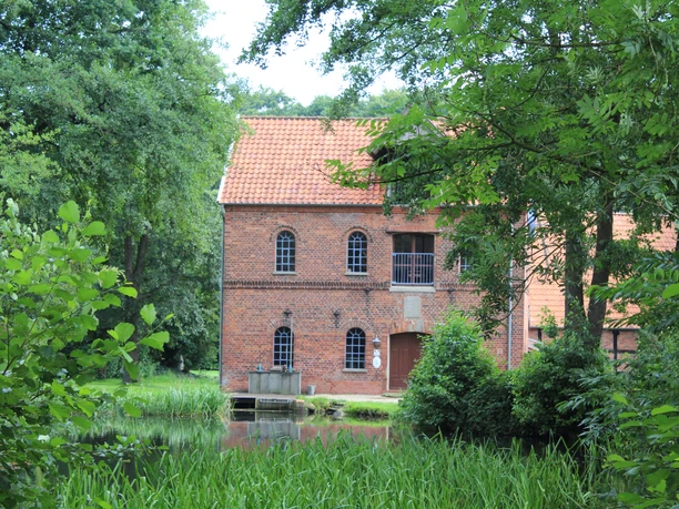 Wassermühle Süstedt Historische Wassermühle aus rotem Backstein umgeben von grüner Vegetation am Wasser in ländlicher Umgebung.
