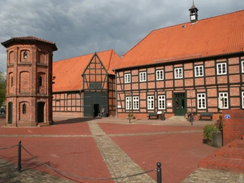 Backsteinbau mit rotem Dach, zweistöckiger Turm im Vordergrund, historische Fachwerkkonstruktion.