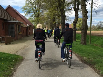 Wolfstour Nord Eine Gruppe von Radfahrern auf einer schmalen Landstraße, flankiert von Bäumen und Ziegellandhäusern.A group of cyclists on a narrow country road, flanked by trees and brick country houses.En gruppe cyklister på en smal landevej, flankeret af træer og murstenshuse.Een groep fietsers op een smalle landweg, geflankeerd door bomen en bakstenen landhuizen.
