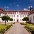 Historische Klosteranlage mit symmetrischen Gebäuden, gepflegtem Hof und Baumreihe unter blauem Himmel.