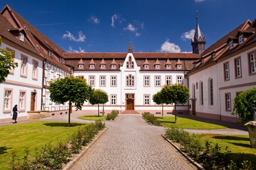 Kloster Brede Historische Klosteranlage mit symmetrischen Gebäuden, gepflegtem Hof und Baumreihe unter blauem Himmel.
