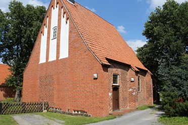 Red brick church with a steep roof and white field orders, surrounded by green trees.