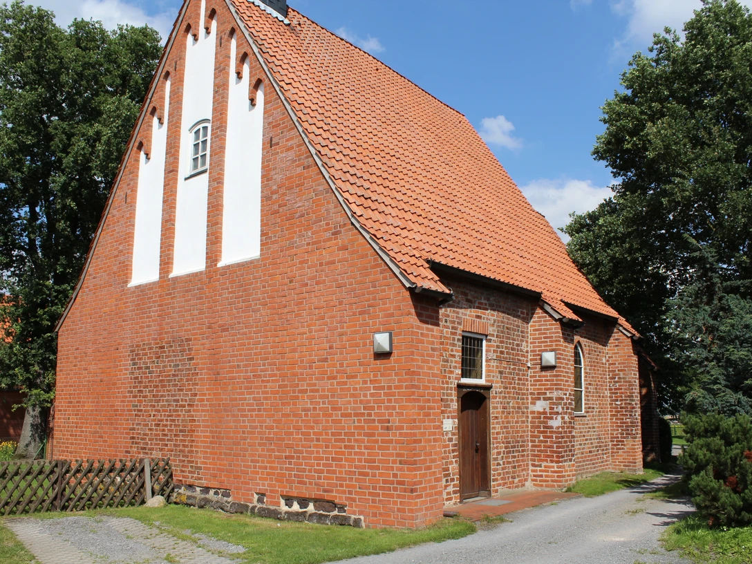 Rote Backsteinkirche mit steilem Dach und weißen Feldordnungen, umgeben von grünen Bäumen.