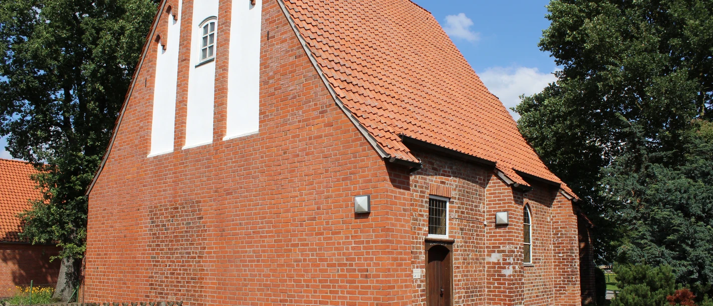 Red brick church with a steep roof and white field orders, surrounded by green trees.
