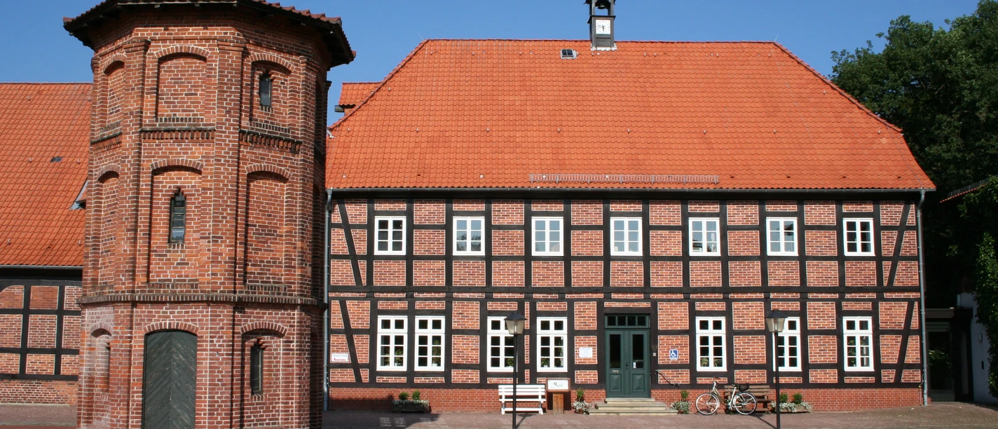 Historisches Fachwerkgebäude mit rotem Ziegeldach und Turmanbau auf einem gepflasterten Platz.