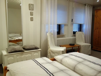 Ferienwohnung Kuhnert - Schlafzimmer Helles Schlafzimmer mit Doppelbett, zwei bequemen Sesseln, Holzmöbeln und einem großen Spiegel.