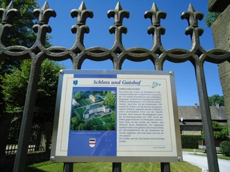 Übersicht Schloss Fürstenberg Informationstafel vor einem Eisengitter, dahinter Schlossbauten und blauer Himmel sichtbar.